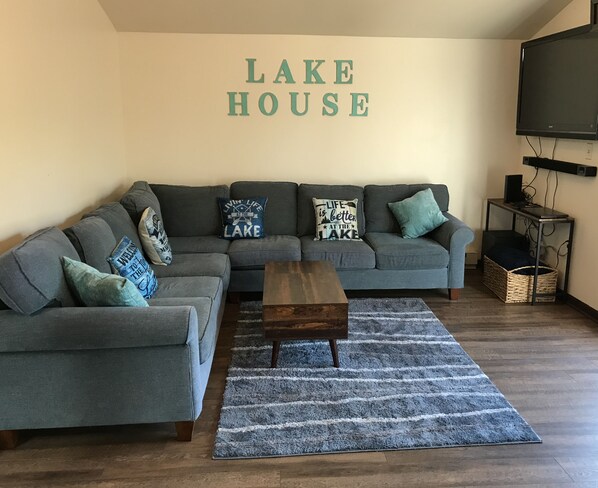 Una sala de estar con un sofá seccional gris, una mesa de centro de madera y un tapete azul. En la pared, un letrero que dice "LAKE HOUSE".