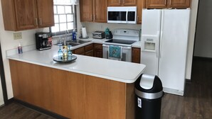 A kitchen with wooden cabinets, a white countertop, a stainless steel sink, a microwave, a stove, a refrigerator, and a trash can. - Lake House w/ Pool and King Bed, Fishing - Dogs OK (Hedgesville)