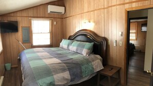A bedroom with a wooden paneled ceiling, a bed with a checkered bedspread, a bedside table with a remote control, and a wall-mounted TV. - Lake House w/ Pool and King Bed, Fishing - Dogs OK (Hedgesville)