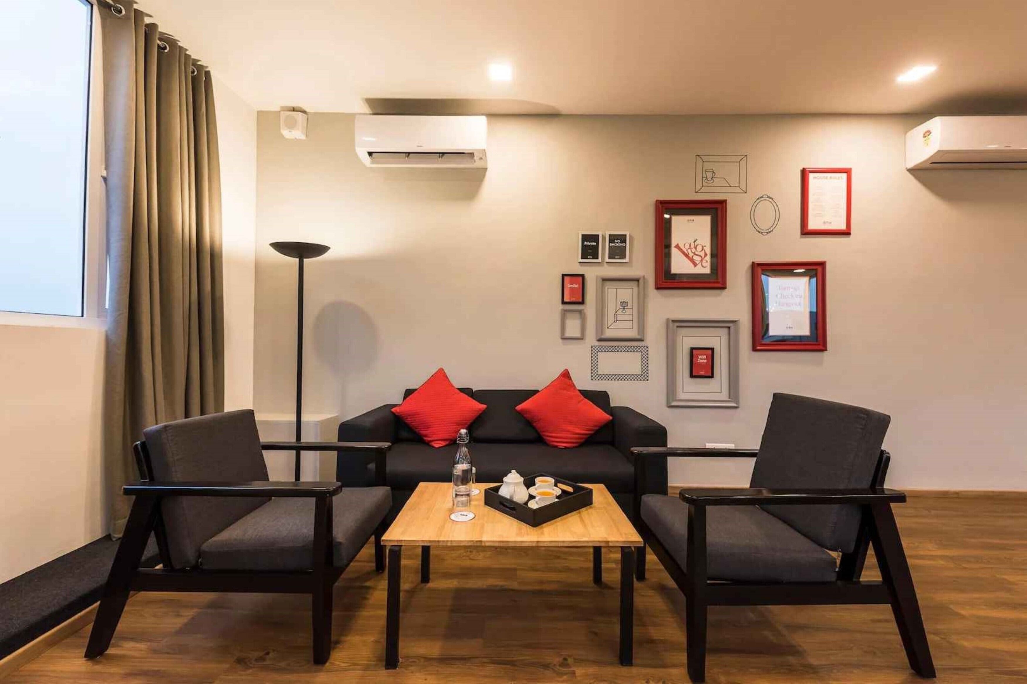 Una sala de estar moderna con un sofá negro, dos sillones, una mesa de centro de madera y una lámpara de pie. Hay cuadros enmarcados en la pared y una unidad de aire acondicionado arriba.