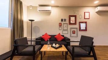 Una sala de estar moderna con un sofá negro, dos sillones, una mesa de centro de madera y una lámpara de pie. Hay cuadros enmarcados en la pared y una unidad de aire acondicionado arriba.