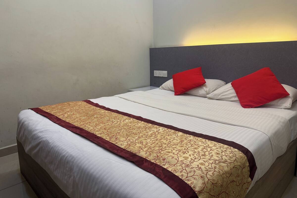 A neatly made bed with a patterned bedspread and two red pillows, a plain wall, and a light switch.