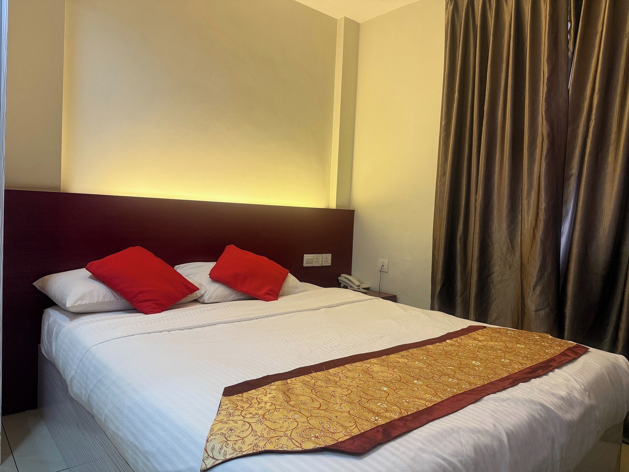 Une chambre d’hôtel avec un lit, des oreillers rouges, un couvre-lit blanc et un chemin de table décoratif aux motifs dorés et rouges.