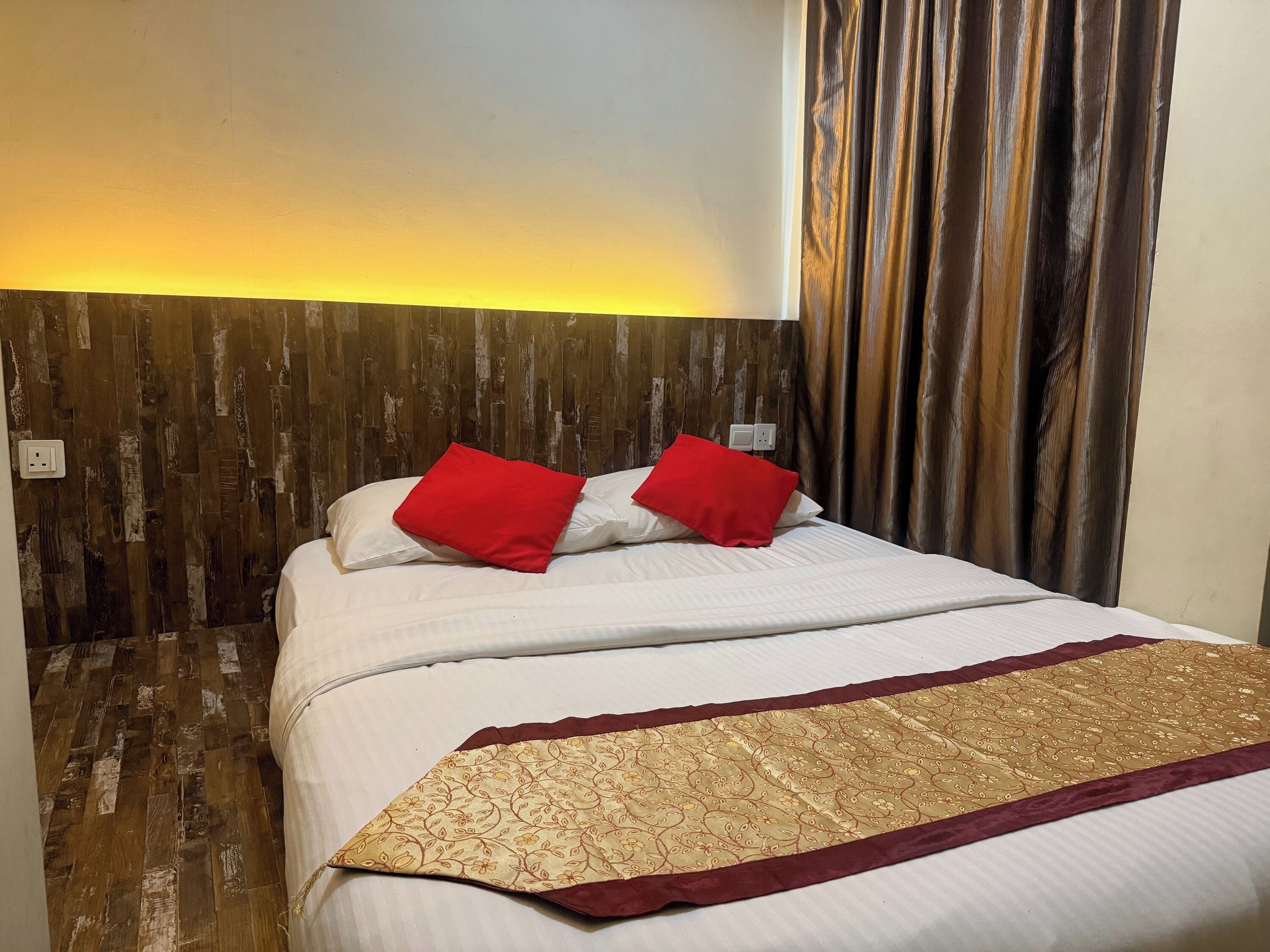 Habitación de hotel con cabecera de madera, cama con ropa de cama blanca y almohadas rojas, una manta con estampado y una cortina.