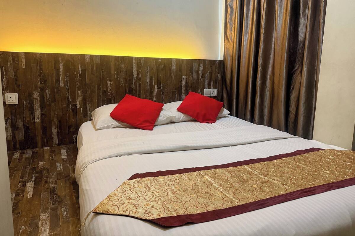 A hotel room with a wooden headboard, a bed with white bedding and red pillows, and a patterned throw blanket.