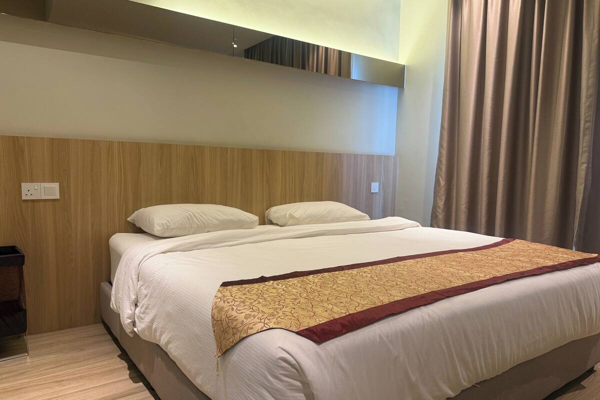 A hotel room with a bed, a wooden headboard, a wall-mounted light, and a window with curtains.