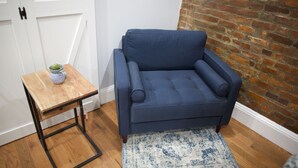 Ein blauer Sessel mit einem hölzernen Beistelltisch und einem blauen Teppich in einem Raum mit Backsteinwand und Holzboden.