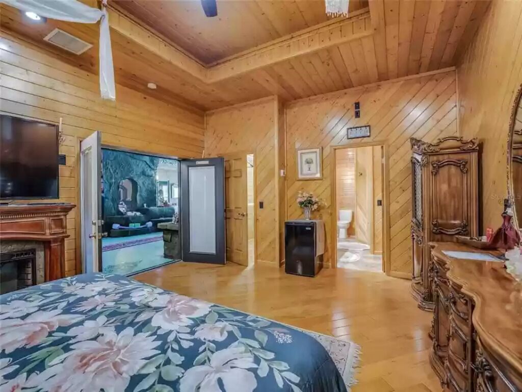 A wooden-paneled room with a bed, a television, a fireplace, and a hallway leading to another room.