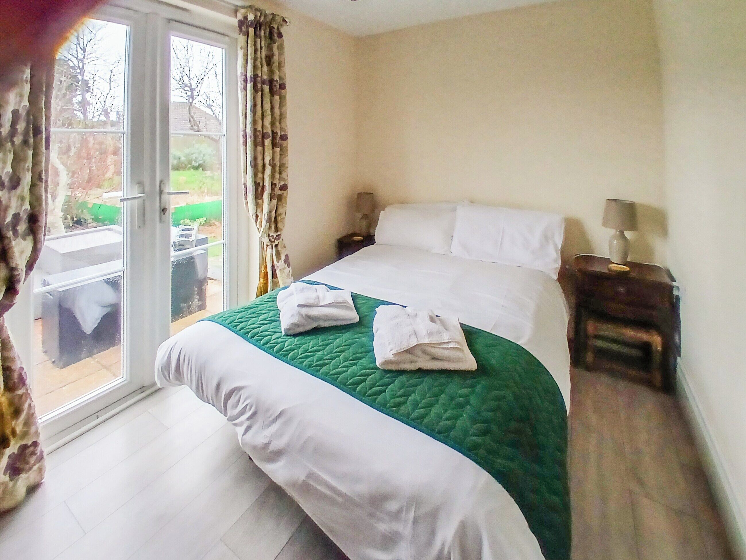 A hotel room with a bed, a nightstand, a lamp, and a view of the outside.