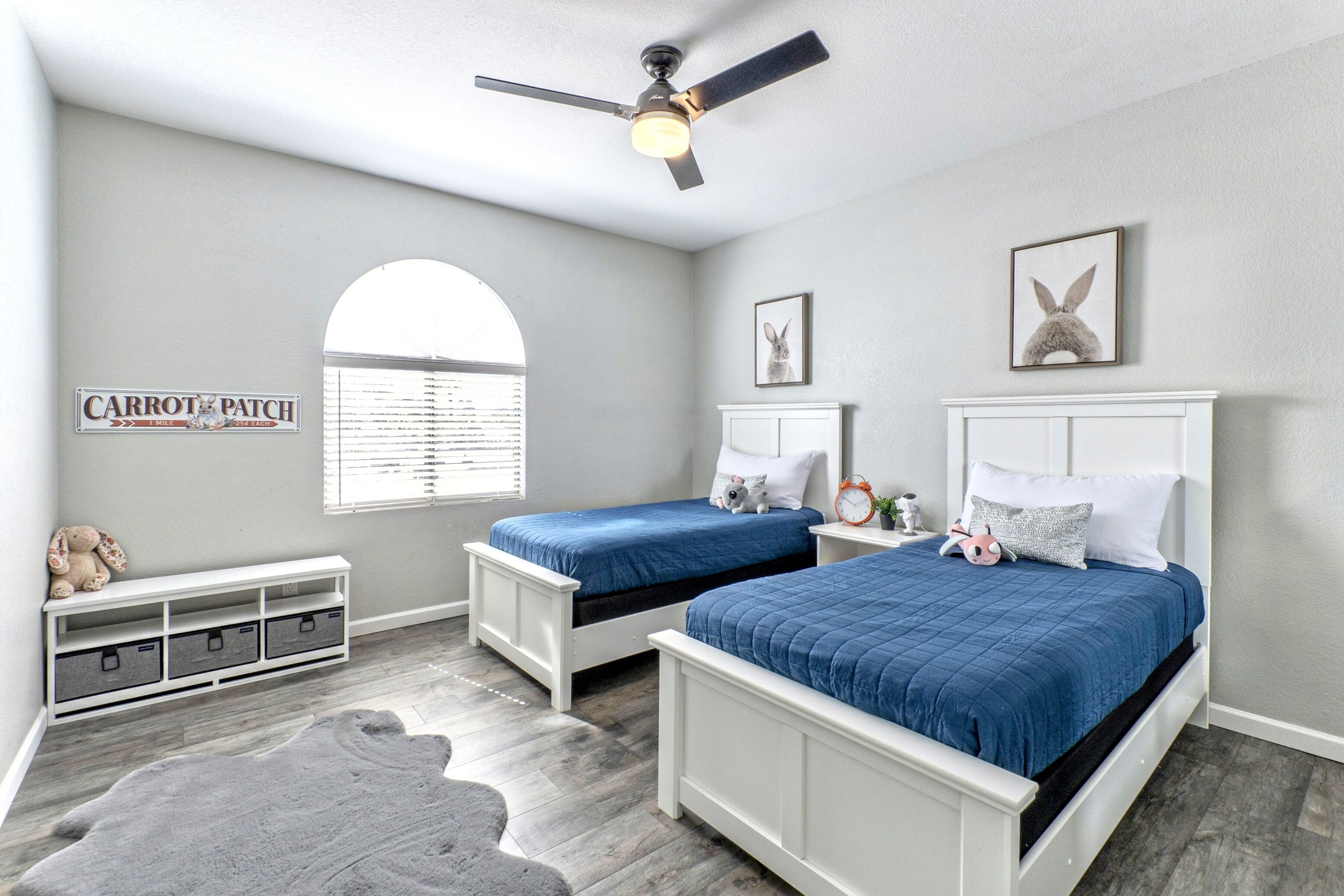 A bedroom with two beds, a ceiling fan, a window with blinds, a storage unit, and a rug.