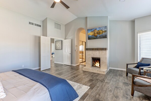 Une chambre spacieuse avec un grand lit, une cheminée, un coin salon avec un fauteuil et une table, et une porte donnant sur une autre pièce.