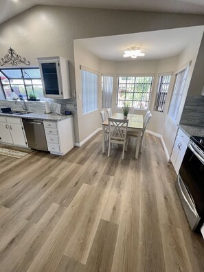 A modern kitchen with a dining area, wooden flooring, white cabinetry, and a window with blinds.