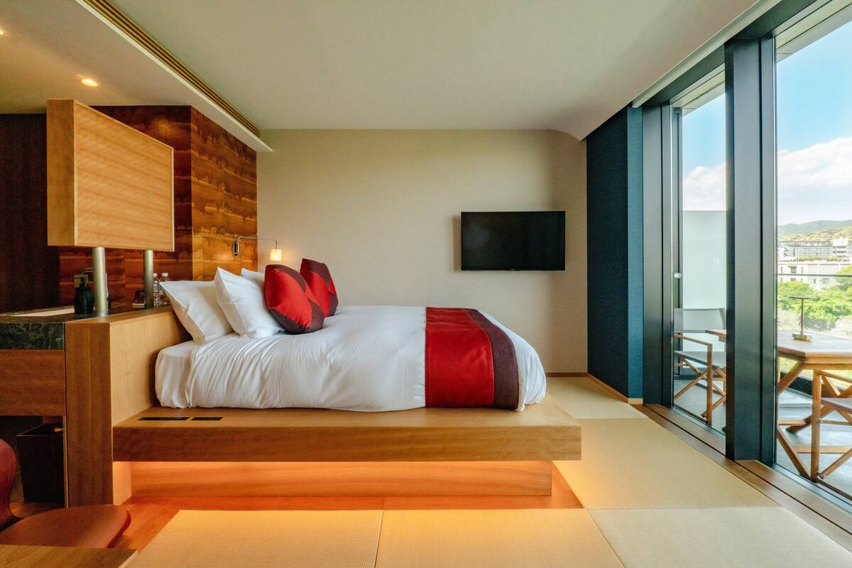 A modern hotel room with a large bed, a flat-screen TV, and a view of the city through large windows.