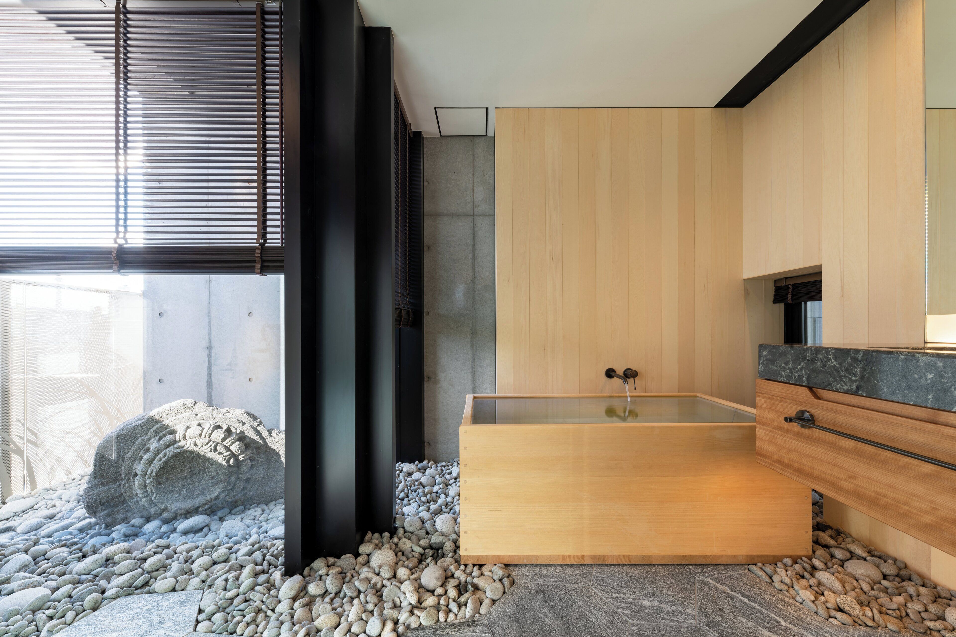 A modern bathroom with a wooden bathtub, a stone wall, and a large window.