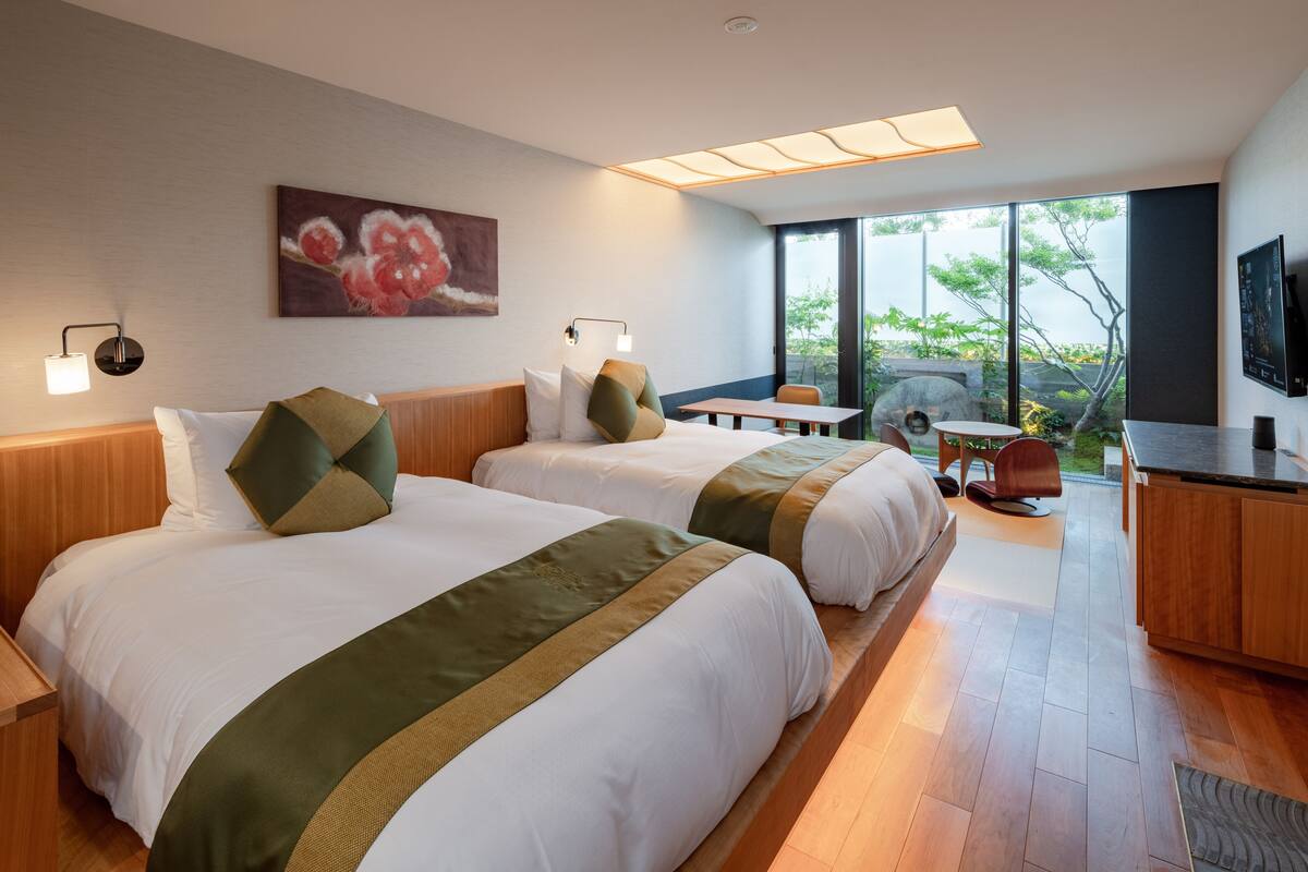 A hotel room with two beds, a desk, a chair, a TV, and a large window with a view of greenery.