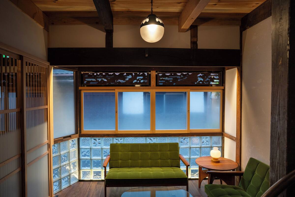 A room with a green sofa, a small table with a lamp, and a view through a large window with wooden frames.