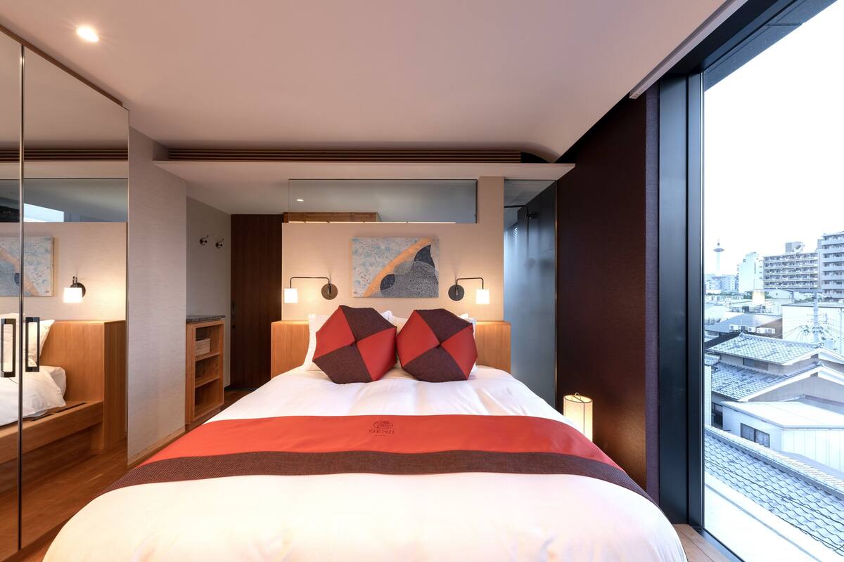A modern hotel room with a large bed, a view of the city, and a wooden wardrobe.