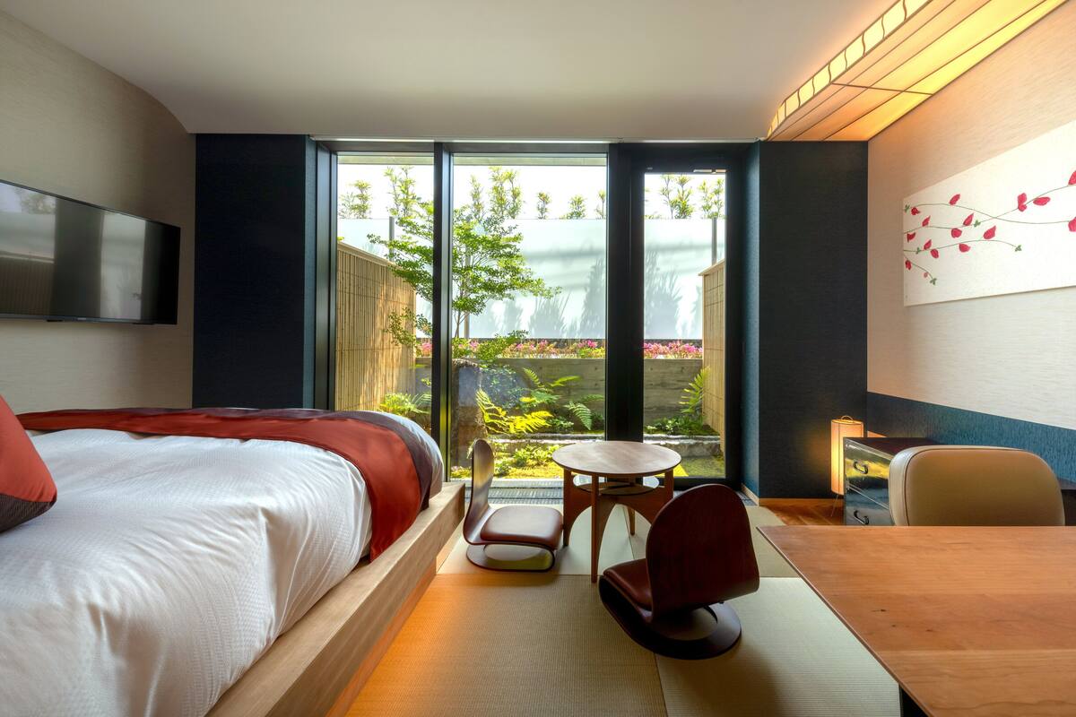 A hotel room with a bed, a desk, a chair, a TV, and a view of a garden through large windows.