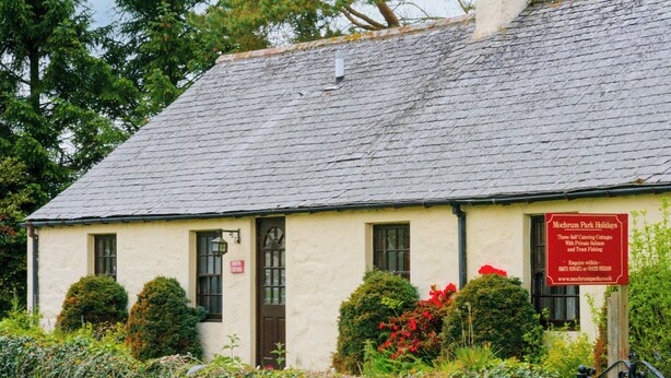 Une maison de campagne traditionnelle, avec un toit en pierre, sur laquelle est apposée une enseigne indiquant qu’elle propose des locations de vacances avec cuisine et un accès à une pêche privée.