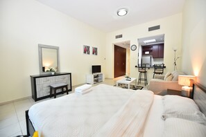 A hotel room with a large bed, a desk with a computer, a TV, and a kitchenette in the background.