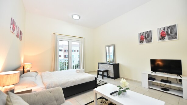 A modern hotel room with a bed, a desk, a TV, and a window with a view of buildings. - KEN - Spectacular Furnished Studio (Dubai)