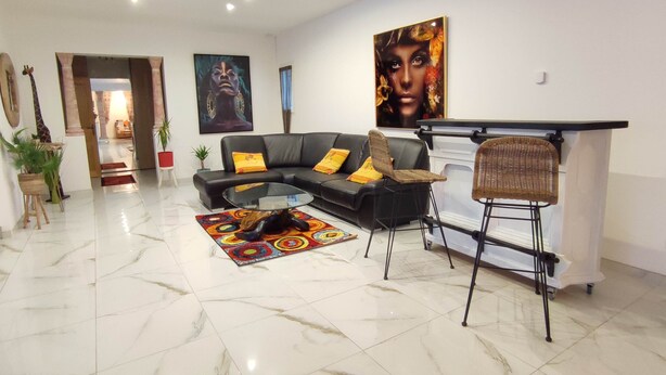 A modern living room with a black sectional sofa, a glass coffee table, a colorful rug, and a wicker chair. There are two large paintings on the walls and a doorway leading to another room. - Villa Carthage confort & vue piscine XL / Downhill stylish Villa view & XL pool (Calignac)