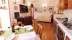 Uma sala de jantar com mesa posta para dois, um sofá e uma cozinha com armários de madeira e fogão.