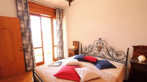 Quarto com cama, piso de madeira, cortinas, porta de madeira, mesa de cabeceira e um criado-mudo.