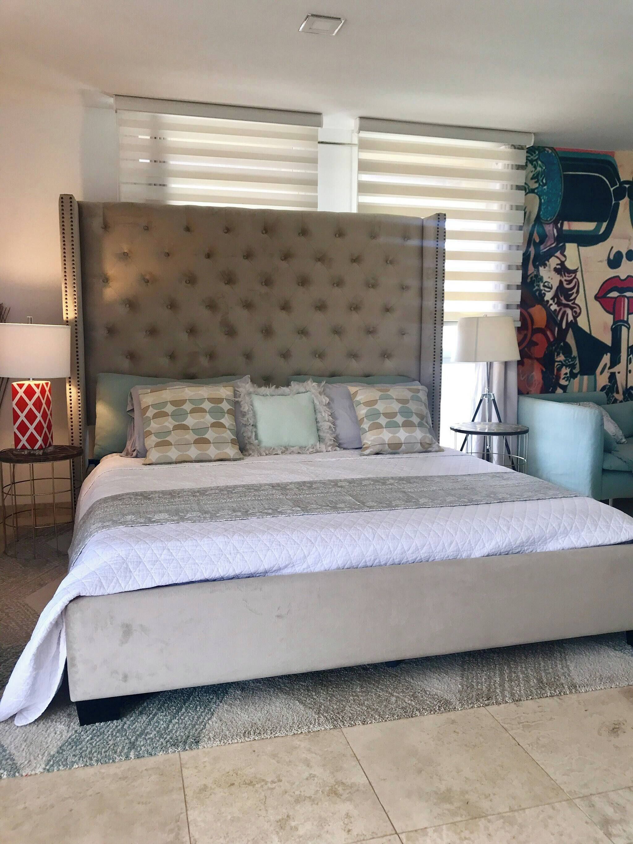 Un dormitorio con una cama grande, un cabecero, una mesita de noche, una lámpara y una pared con un mural.
