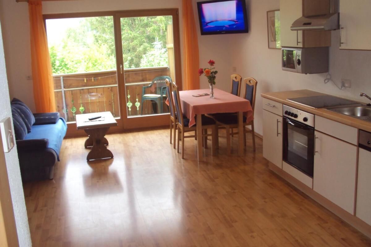 A compact kitchen with a dining area, a TV mounted above the counter, and a balcony with a view of greenery.
