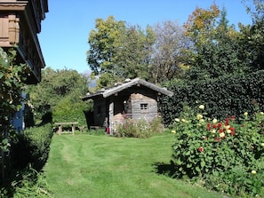 Une cabane en bois rustique, dotée d’un toit en pente, entourée d’un jardin bien entretenu où poussent diverses plantes et fleurs.