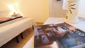 Uma cama bem arrumada com um edredom branco e um travesseiro, uma luminária de cabeceira e uma revista chamada “SALENTO Style” sobre uma mesa.