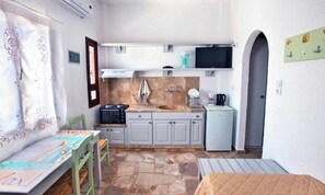 A compact kitchen with a microwave, sink, and small refrigerator. There is a dining table with chairs and a bed in the corner.