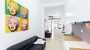 Una sala de estar moderna con un sofá negro, un cuadro de arte pop colorido, una cocina con encimera de madera y una estantería al fondo.
