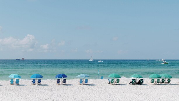 Una playa con filas de sillas y sombrillas, cielo despejado y barcos en el agua.