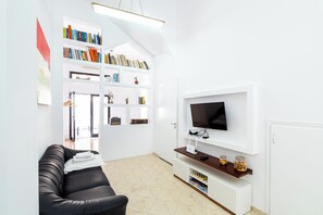 A modern living room with a black sofa, a flat-screen TV, white shelving with books and decorative items, and a tiled floor.