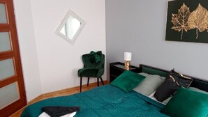 A bedroom with a teal bedspread, green pillows, a small chair, a bedside table with a lamp, and a framed leaf artwork on the wall. - Apartment in the Center - na Ratuszu (Jelenia Gora)