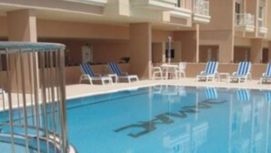 Una piscina con una valla de seguridad metálica, sillas de descanso y un edificio con balcones al fondo.