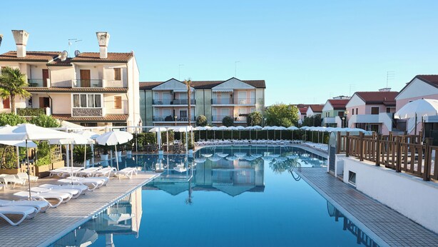 Une piscine entourée de bâtiments dotés de balcons, avec des chaises longues et des parasols, le tout sous un ciel dégagé.
