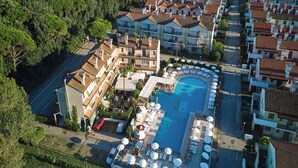 Vue aérienne d’un complexe hôtelier doté d’une piscine entourée de chaises longues et de parasols, situé à proximité d’immeubles résidentiels.