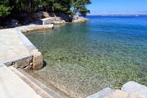 Zona costera despejada con un sendero de piedra que lleva al agua, rodeada de árboles y una vista distante del mar.