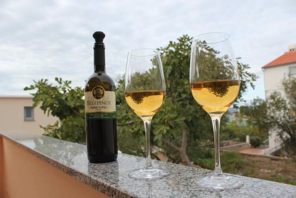 En flaske Bel Pinot vin og to glas på en balkon med udsigt til træer og bygninger.