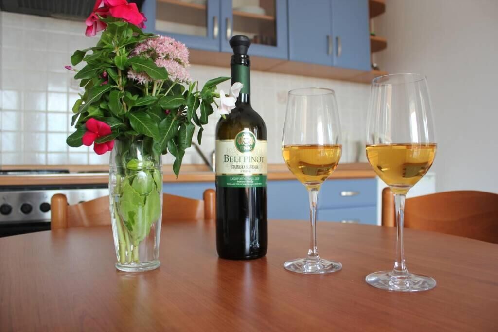 Una botella de vino Belli Pinot, dos copas de vino y un jarrón con flores sobre una mesa de madera en una cocina.