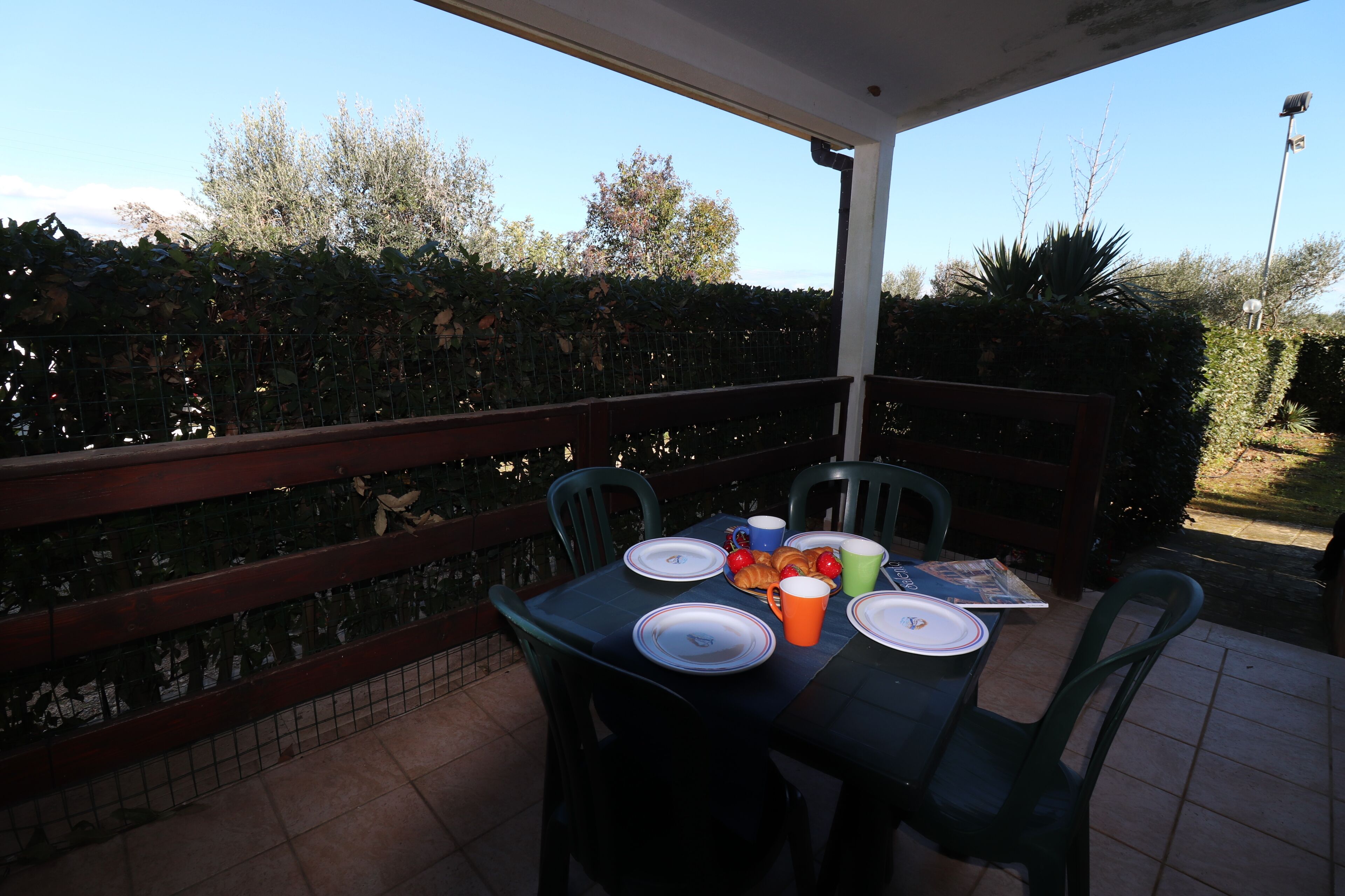 Eine überdachte Terrasse mit Tisch für zwei Personen, gedeckt mit Tellern, Tassen und einem Croissant.