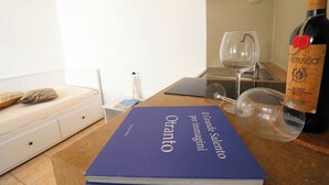 Une pièce comprenant un lit, une table avec une bouteille de vin et des verres, et un livre intitulé « Otrante ».