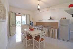 A kitchen with a dining table, chairs, a refrigerator, a sink, and a window with a view of greenery.