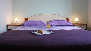 Un lit recouvert d’une couvre-lit violette, deux coussins à motifs, un plateau avec un croissant et un verre de jus d’orange, et deux lampes de chevet.