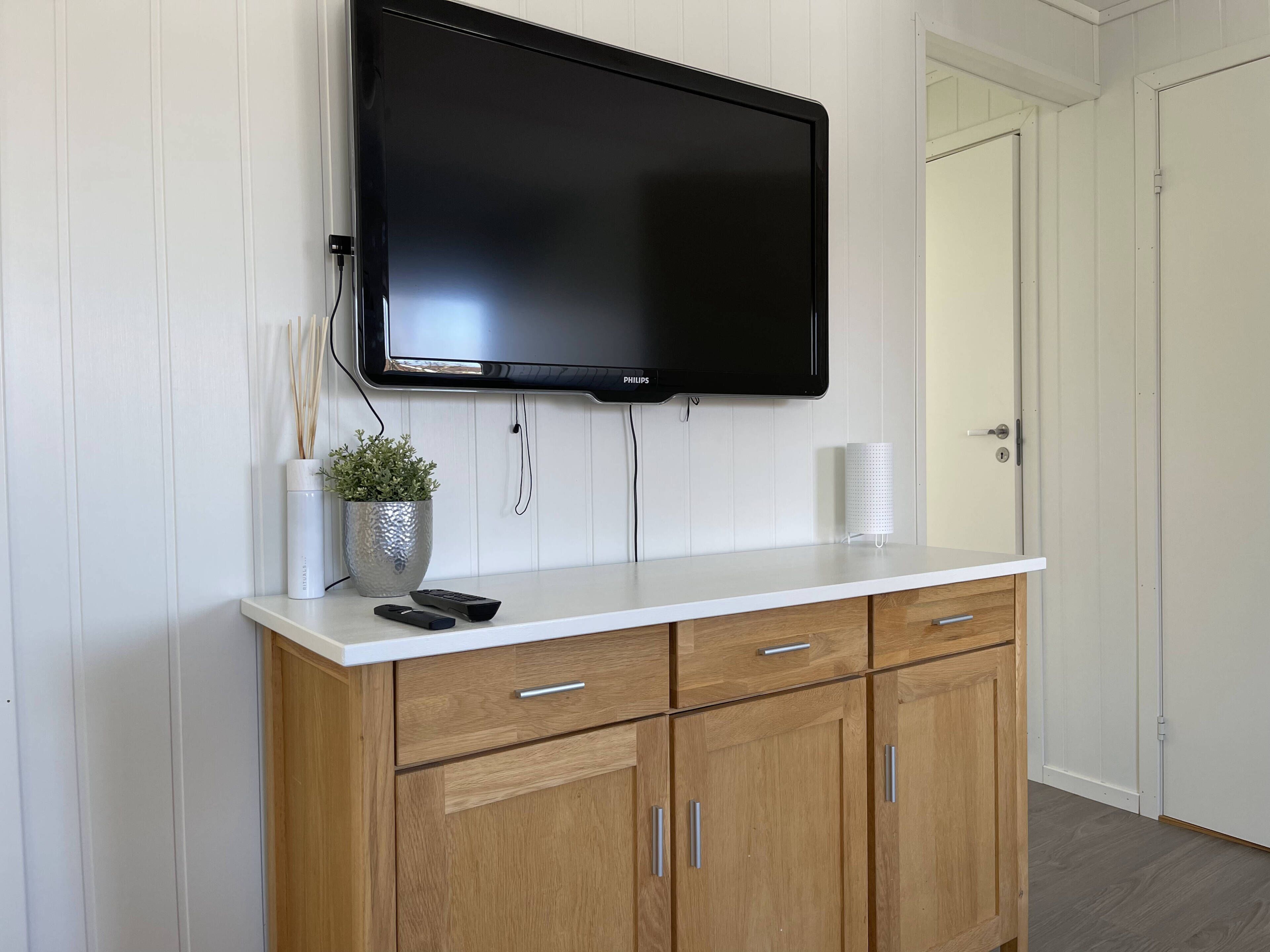 A modern room with a flat-screen TV mounted on a white wall, wooden cabinets below, a small potted plant, and a white door.