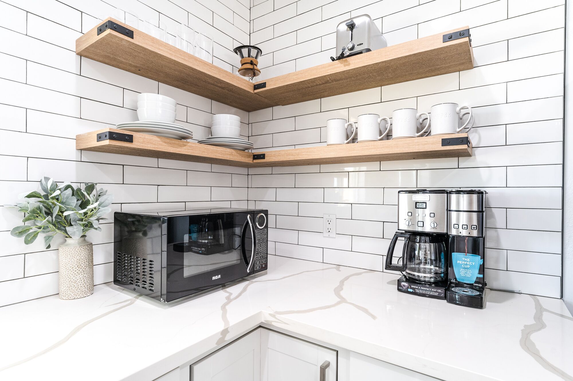 Una cocina moderna con azulejos blancos tipo metro, estantes de madera, un microondas y una cafetera.