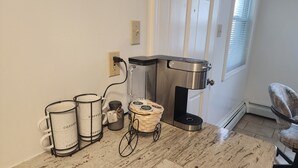 Una estación de café con una cafetera, un cesto de cápsulas de café y tres tazas sobre un suelo de baldosas.